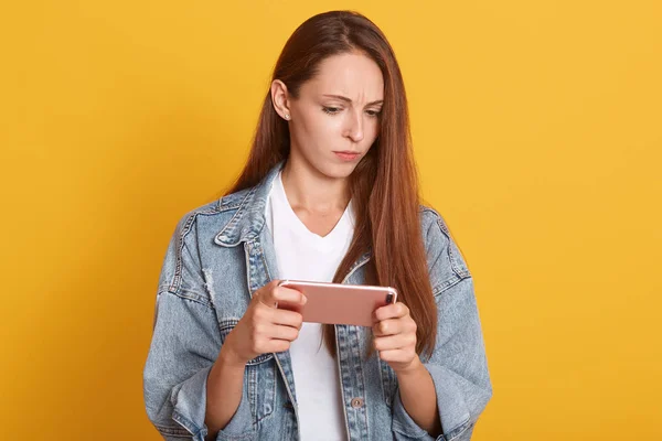 Porträt einer jungen Frau in Jeans lässige Kleidung mit Handy und Blick auf seinen Bildschirm isoliert auf der gelben Wand im Studio, mit missbilligendem Gesichtsausdruck, versteht nicht, was sieht. — Stockfoto