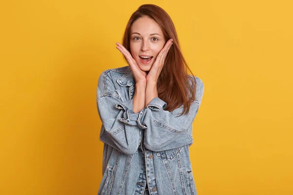 Potret dekat gadis muda telah terkejut ekspresi wajah dengan tangan di pipinya. Wanita muda cantik yang memegang mulutnya membuka, mengenakan jaket denim berpose terisolasi dengan latar belakang kuning . — Stok Foto