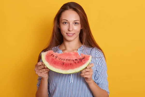Horizontal shot of young woman with piece of watermelon in hands, female wearing striped shirt and having long brown hair, looking smiling at camera, model posing isolated over pink studio background.