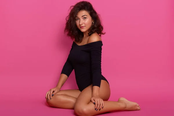 Indoor shot of young slim lady with dark wavy hair dressed black combi dress, poses with bared shoulders, sitting on the floor over pink studio background, looking at camera with passionate expression — Stock Photo, Image