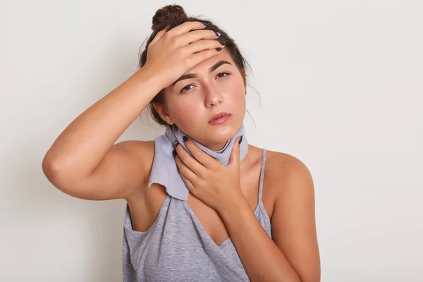 Woman puts hands on head and on throat, having scarf around neck, has high tempereature, lady wearing gray t shirt, posing isolated over white studio background. Healthy and illness concept.