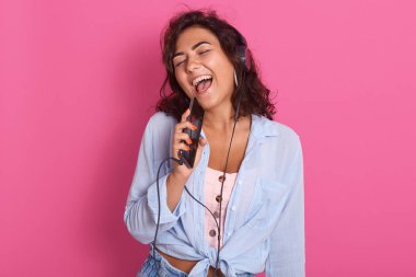 Genç kız dinleme ve müzik zevk, mikrofon olarak hücresel tutarak, mp3 çalar olarak akıllı telefon kullanımı, başının üstünde kulaklık var, şık giyim giyiyor, pembe arka plan üzerinde izole.