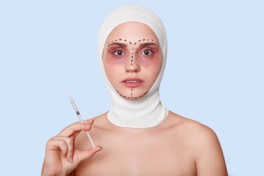 Siyah düzeltme işaretleri ile kadının yüzüne yakın atttraktif kadın tutan şırınga ön görünümü, mavi stüdyo arka plan üzerinde izole poz modeli, bared omuzlar ve başında bandajlar ile kadın.