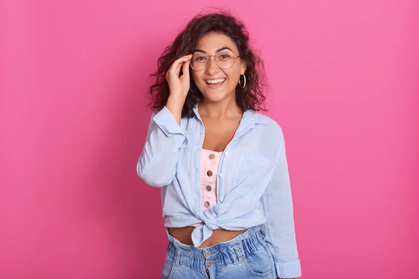 Indoor shot de hermosa chica con gafas sobre fondo rosa, señora con top rosa, camisa azul y jeans, mirando sonriendo directamente a la cámara, mantiene la mano en sus gafas, tiene expresión feliz . —  Fotos de Stock