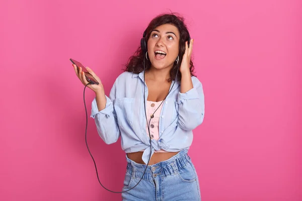 Bella e felice giovane donna ascoltare musica con le cuffie, isolato su sfondo rosa stuio, attraente femmina indossa abbigliamento elegante tenendo smartphone in mano, divertirsi a casa . — Foto Stock