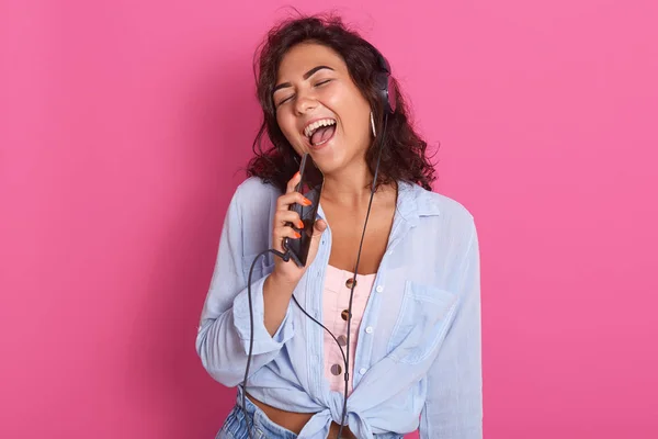 Young girl listening and enjoying music, holding cellular as microphone, use smart phone as mp3 player, has earphones on top of her head, wearing stylish clothing, isolated on rosy background. — Stock Photo, Image