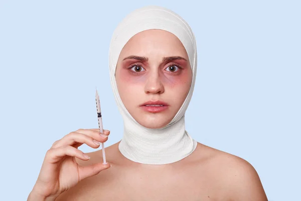 Фото молодої жінки, що тримає шприц для проведення процедури омолодження обличчя, жінки після косметичної операції, мають синці навколо очей та медичну пов'язку, ізольовані на синьому студійному фоні . — стокове фото