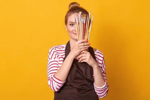 Horizontal shot of girl artist holds brushes in hands and hides behind it, lady dresses stripes casual shirt and brown apron, blonde woman with bunch, posing isolated over yellow studio background.