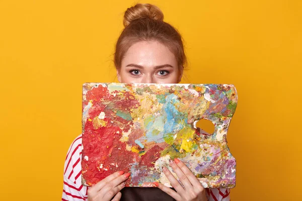 Captura de estudio de la atractiva joven artista sosteniendo y escondiéndose detrás de la paleta de colores o sus obras de arte, mirando a la cámara, adorable mujer con camisa a rayas, posando aislada sobre un fondo amarillo . — Foto de Stock