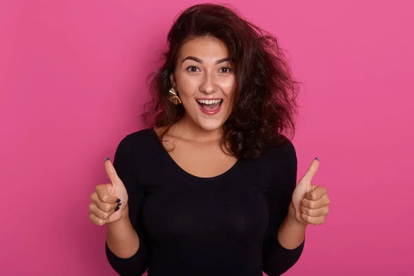 Fronte ritratto di bella giovane donna dando pollici in piedi isolato su sfondo rosato, modello caucasico sorridente felice, indossando camicia nera, con i capelli scuri ondulati. Persone emozioni concetto — Foto Stock