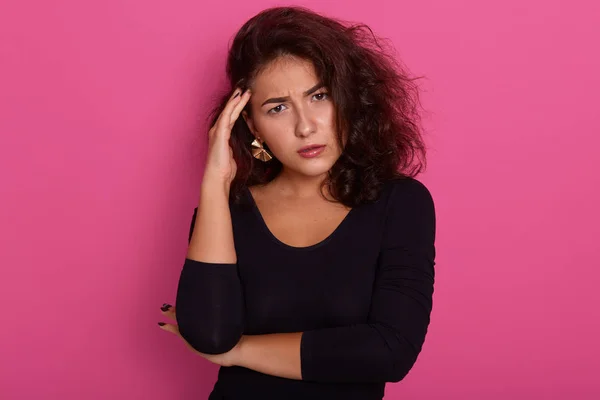 Mujer pensativa que tiene el pelo ondulado oscuro sosteniendo su dedo índice en el templo, chica adorable mirando tristemente a la cámara, posando aislado sobre la pared de hormigón rosa del estudio. Mujer pensativa con dolor de cabeza . — Foto de Stock