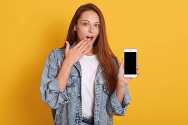 Şok etekli beyaz kız cep telefonunun boş ekranında işaret parmağıyla işaret ederken sarı arka plan üzerinde izole dururken, beyaz gömlek ve kot ceket giyen kadın, açık ağızla poz verdi.