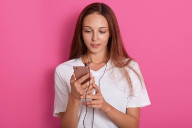 Genç kadın modern cep telefonunu kullanarak, parmakla ekrana dokunmadan, pembe arka plan üzerinde poz, beyaz tshirt giyen, güzel staright saçsahip, müzik dinleme, evde rahatlatıcı