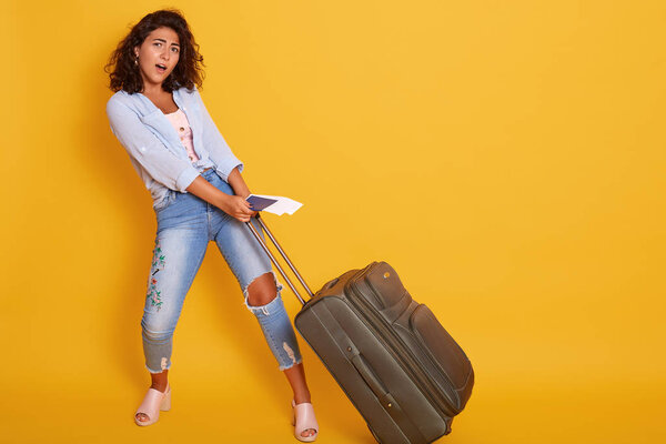 Studio shot of beautiful girl carries heavy suitcase with documents and flight tickets in hand, posing with opened mouth, looks tired, isolated over yellow background. Traveling and lifestyle concept.