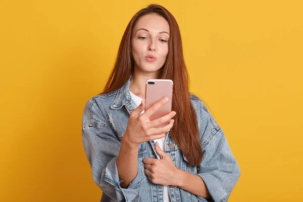 Image of young woman posing for funny selfie, making kiss gesture, looking at camera on her mobile phone, charming lady wearing fashionable denim jacket, posing isolated over yellow studio background. — Stock Photo, Image