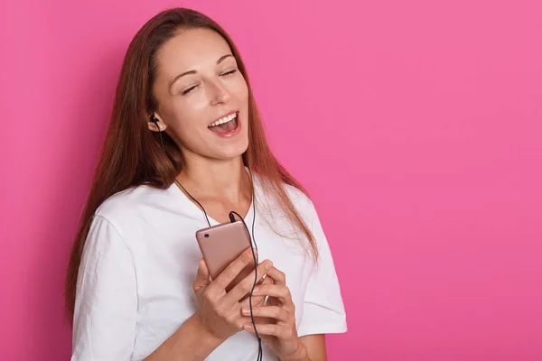 Kép boldog gondtalan fiatal nő táncol és hallgat zenét okostelefon több mint rózsaszín háttér, hölgy visel fehér inget, gazdaság mobiltelefonján. Hirdetés vagy promóciós szöveg másolása. — Stock Fotó