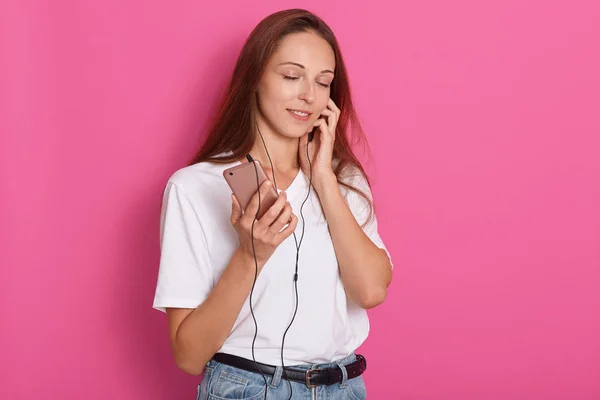 Portrait of beautiful girl in headphones listening to music with closed eyes, standing over pink studio background, wearing white casual t shirt, relaxes with favourite songs. Copy space for promotion — Stock Photo, Image