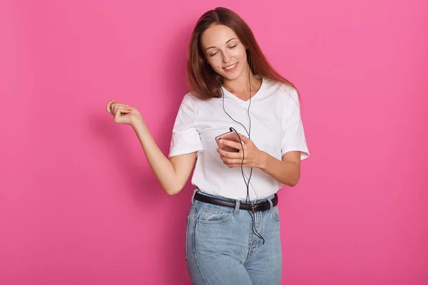 Kvinna dansar med hörlurar Lyssna på musik via Smart Phone. Lekfull glad leende ung kaukasiska kvinna isolerad över Rosen bakgrund, attraktiv dam spenderar ledig tid. Människor Concept. — Stockfoto
