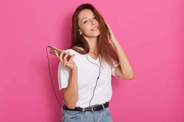 Studio shot of young woman listening to music with earphones, holding smartphone in hands, looking at camera, ralaxing at home, wearing casual clothing, having pleasant look. Free time concept. — Stock Photo, Image