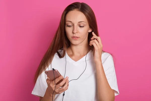 Horizontal shot of dark haired hipster girl listening to audio track with thoughtful facial expression, has ear phones and holding smart phone in hands, isolated on pink background. Lifestyle concept.