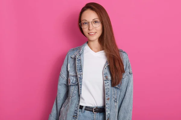 Portrait of happy woman dressed in denim eyewear, jacket and white casual t shirt, poses against pink wall with free space asides, looking directly at camera with nice smile, being in good mood . — Stok Foto