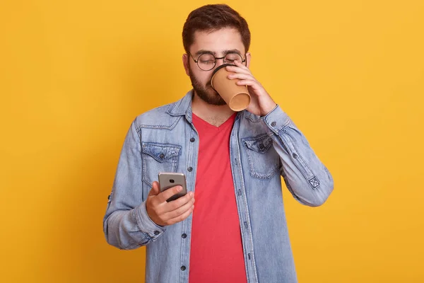 Ελκυστικός νεαρός άντρας πίνοντας καφέ ή τσάι, κρατώντας το έξυπνο τηλέφωνο σε άλλο χέρι και κοιτάζοντας την οθόνη του, φορώντας κόκκινο casual πουκάμισο και ντένιμ μπουφάν. Έννοια της τεχνολογίας και των ανθρώπων. — Φωτογραφία Αρχείου