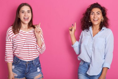 Pembe arka planda işaret eden açık ağızlı iki mutlu gülen kadın görüntü, rahat gömlek ve kot şort giymiş bayanlar, reklam veya promosyon için kopya alanı.