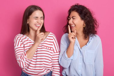 Koyu saçlı, parmaklarını ağızlarında tutan, kasko gömlek giyen, birlikte eğlenen iki genç gülen kız, mutlu yüz ifadelerine sahipler. Gül arka plana karşı konsept stüdyo portresi.