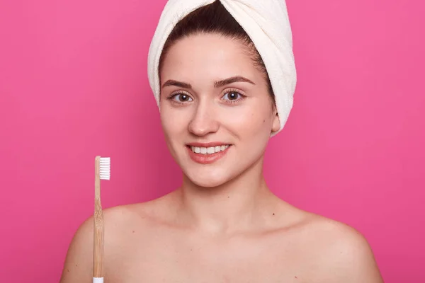 Primer plano retrato de atractiva mujer caucásica sonriente, mira a la cámara, sosteniendo el cepillo de dientes, posando aislado sobre rosado, señora de pie con los hombros desnudos y cepillarse los dientes delante del espejo . — Foto de Stock