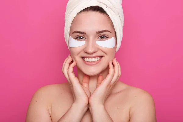 Adult woman stands smiling during spa procedure. Attractive lady with healthy skin posing with patches under eyes and white towel on head, keeps hands nder chin, posing with bared shoulders.