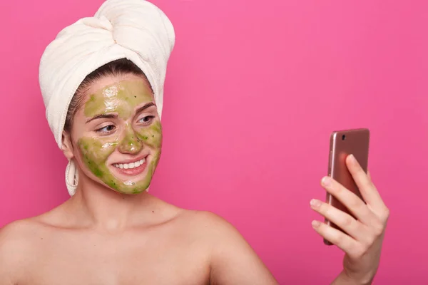 Mooie vrouw met groen masker op gezicht, verpakt witte handdoek, poses half naakt, het nemen van selfie op smartphone in de badkamer, poseren geïsoleerd over roze achtergrond. Cosmetologie en Beauty concept. — Stockfoto