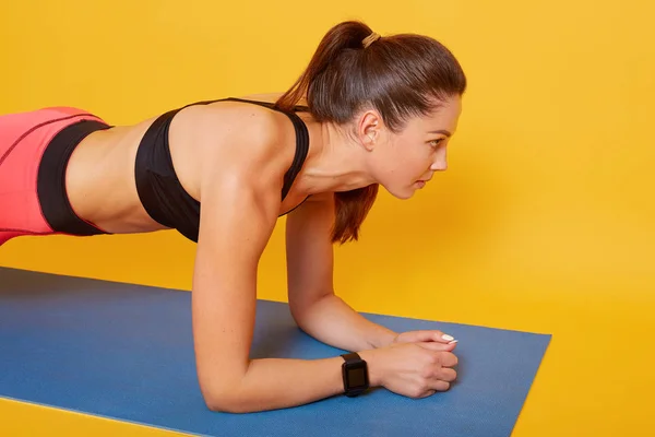 Estudio de tiro de mujer joven haciendo ejercicio, ajuste morena deportiva con cola de caballo haciendo tablón en estera de yoga, vestidos de dama atractiva ropa deportiva, aislado en amarillo. Estilo de vida saludable y concepto deportivo . — Foto de Stock