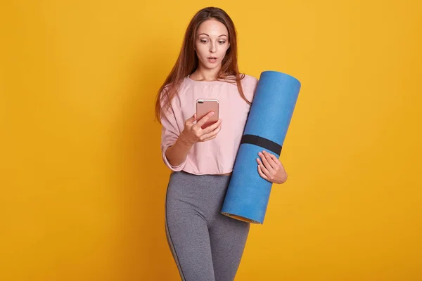 Indoor shot of young yoga instructor stand holding mat, checking mail on smartphone, fit female wearing sportswear use mobile phone, waiting for training in fitness studio, isolated over yellow wall.