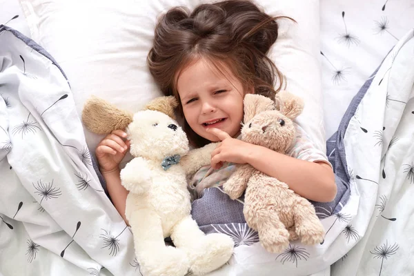 Top view of child crying in bed, sad kid on pillow in bedroom, liitle girl lying on linens with dandelion, upset toddler in pajama lay swith her teddy bears in bed in morning. Childhood concept. — Stock Photo, Image