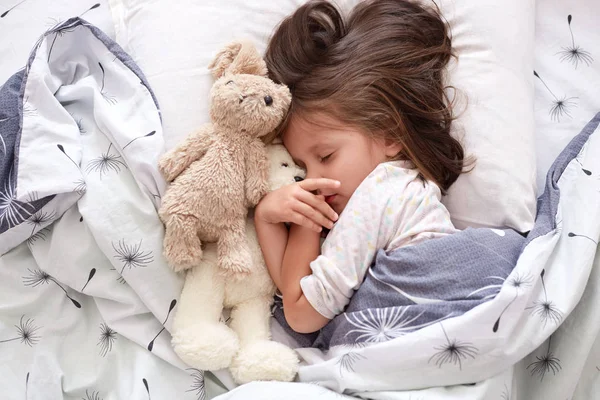 Közelről portré kisgyermek lány szóló vele játék mackó az ágyban, pózol csukott szemmel, miután pihenés kemény nap után, sötét hajú gyermek alszik vele kedvenc játék. Gyermekkori koncepció. — Stock Fotó