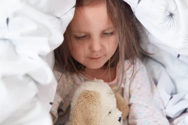 Beltéri lövés imádnivaló kisgyerek lány játszik a mackó az ágyban alatt takaró, bájos gyerek játszik reggel, mielőtt Kinder Garten, baba neki mackó alá kiterjed néz rá a fehér játék. — Stock Fotó