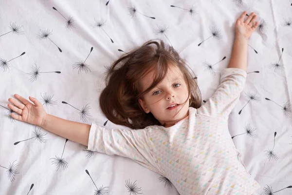Top view of little girl laying in bed and spreading her arms on white cover with dandelion, charming kid with dark hair wearing pajama, looking at camera, relaxing after hard day. Children concept. — Stock Photo, Image