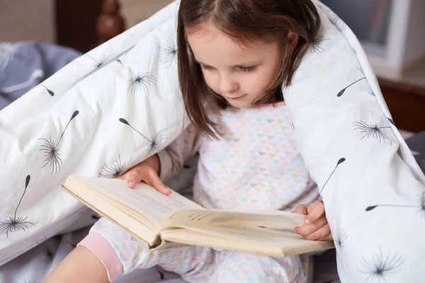Horizontal shot of girl reading under blanket with dandelion, having serious facial expression, looks concentrated, charming toddler holding book in hands, reads fairy tale. Childhood concept. — Stock Photo, Image