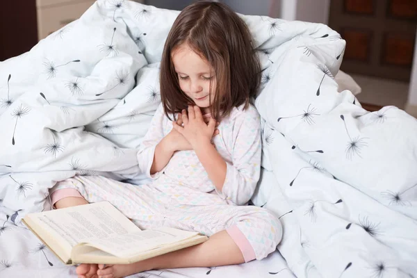 Indoor shot of adorable girl reading book while sitting in bed, cute child looks in book with great interest and keeps hands on chest, looks concentrated, reads with delight. Childhood concept. — Stock Photo, Image