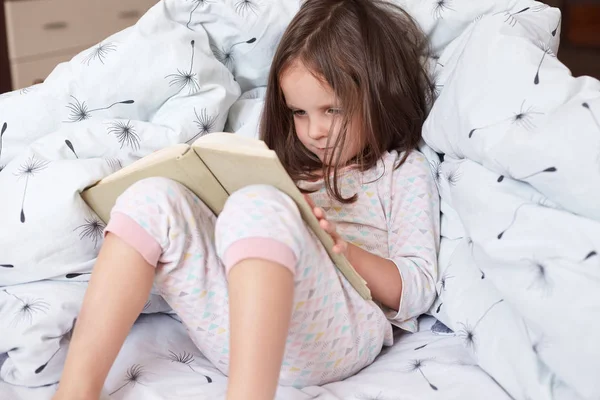 Portrait of adorable child reading book at home while sitting in bed, holding book in hands, looks concentrated, spending morning time with book, cute girl likes fairy tales. Children concept. — Stock Photo, Image