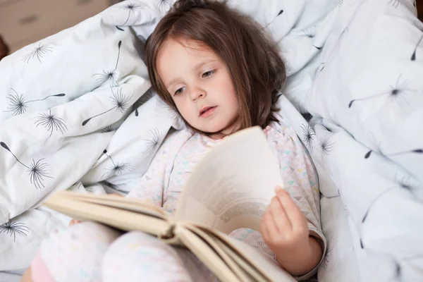 Cute girl reading book siting in her bed and readsinteresting fairy tales, turning pages and finds colourful pictures or interesting stories, charming kid lying on pillows. Childhood concept. — Stock Photo, Image