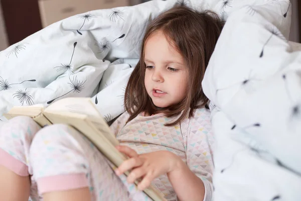 Close up portrait of caucasian baby girlreading book and sitting in bed in her bedroom, baby with concentrated facial expression, preschool child reading fairy tales before go to bed, Children concept — Stock Photo, Image