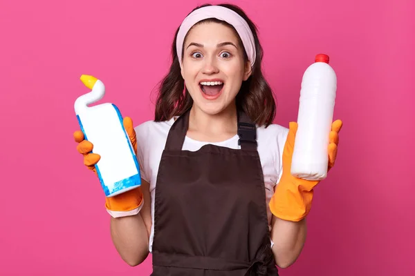 Indoor shot of positive charming young housewife wearing headband, rubber gloves and apron, holding detergents in her hands, having happy facial expression and looks smiling directly at camera. — Stock Photo, Image