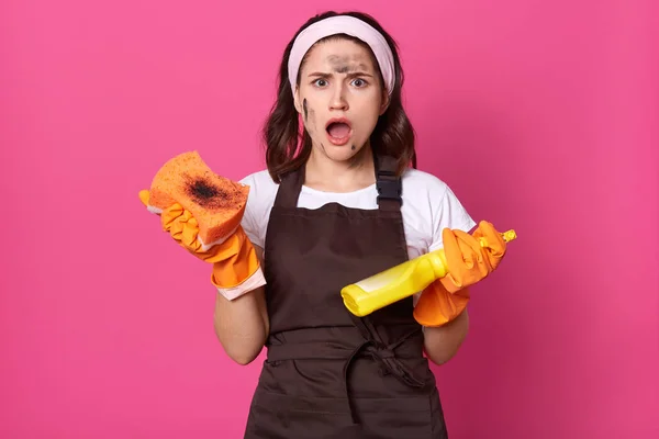 La toma de estudio de una mujer guapa, sosteniendo esponja sucia y detergente en las manos, mantiene la boca abierta, ha sorprendido y conmocionado la expresión facial, cansándose de la casa hendidura. Concepto de servicio doméstico . —  Fotos de Stock