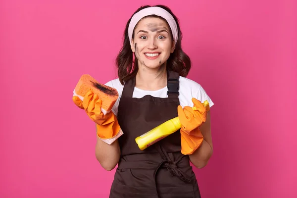 Charming young Caucasian housewife wearing headband, rubber gloves and apron, holding sponge and detergent in hands, looks smiling at camera, being happy to finish cleaning house, hygiene concept. — Stock Photo, Image