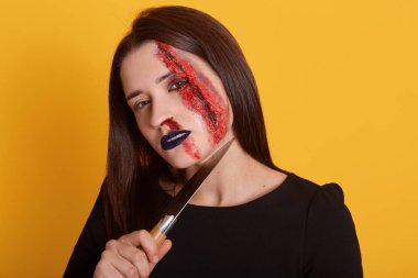 Esmer kadın boynuna yakın bıçak ve yüzünde kanlı yara ile kapalı çekim, doğrudan kameraya bakarak, elbiseler siyah gömlek, sarı stüdyo arka plan üzerinde izole poz. Cadılar Bayramı konsepti.