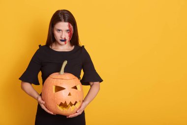 Stüdyoda cadı kadının sarı arka plan üzerinde izole stüdyoda balkabağı tutan, siyah elbise giyen, şöhret korkudolu yüz ifadesi olan vurdu. Kabak baş jack fener ve cadılar bayramı kavramı.