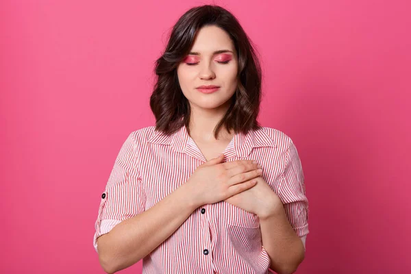 Primer plano retrato de mujer de aspecto amigable, mantiene las manos en el pecho y con los ojos cerrados, se ve con alegría, con camisa casual a rayas, posando aislado sobre fondo rosa. Gente emociones concepto . — Foto de Stock