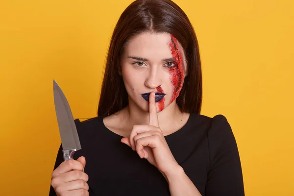 Disparo horizontal de la joven morena con estilo de Halloween maquillaje sangriento, de pie y mirando directamente con el dedo cerca de su boca, muestra silencio, posando sobre fondo amarillo. Concepto Halloween . — Foto de Stock