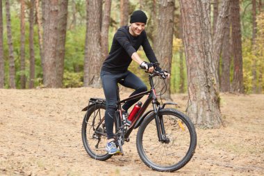 Ormanda gezen, siyah bisiklet giysileri, şapka ve spor ayakkabı giyen, mutlu yüz ifadesine sahip, sportif erkek boş zamanlarını açık havada geçiren, trrees'lerde gezen bisikletçinin pictiure'u.